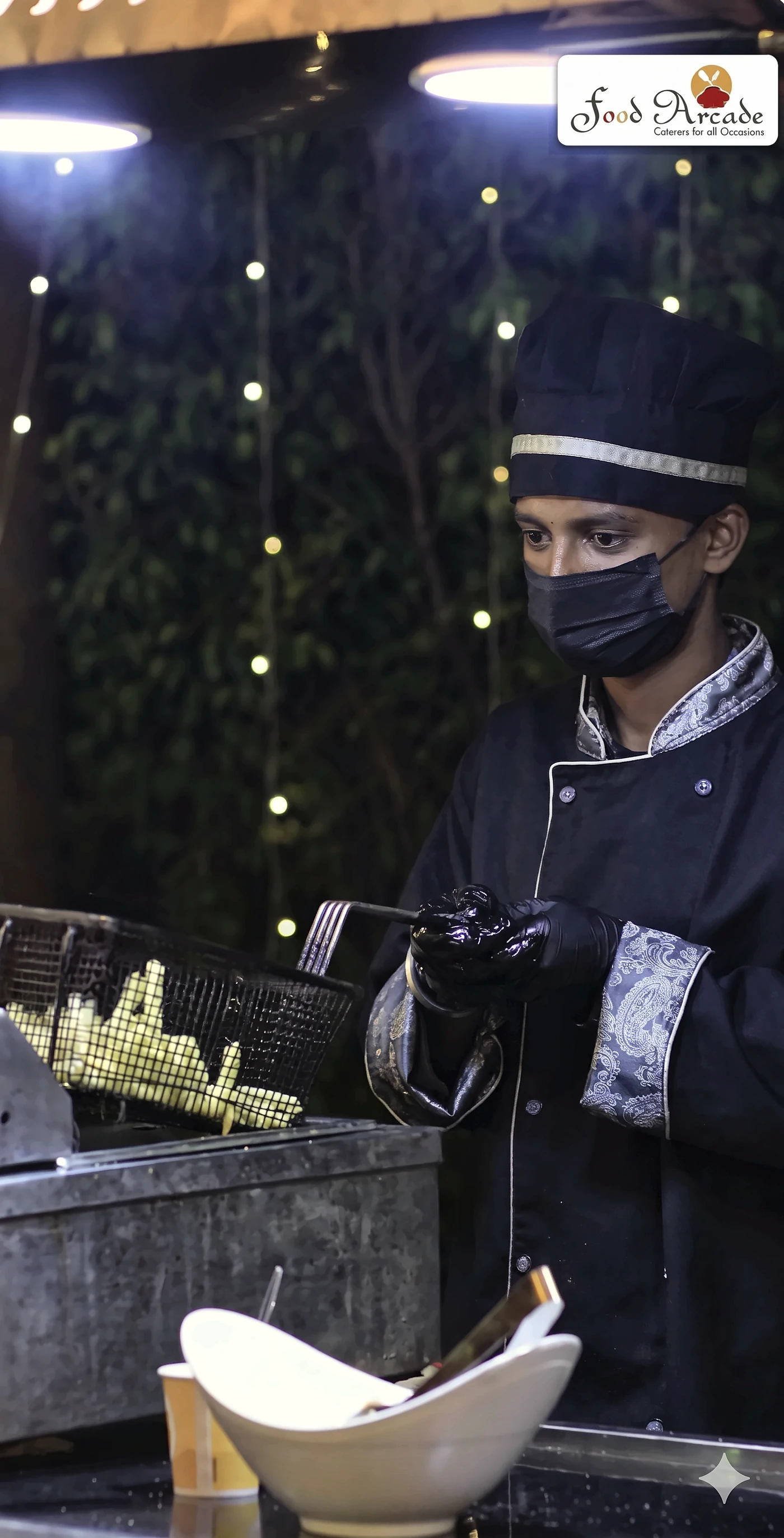 Chef in a black jacket and cap lifting a basket of fries beside a garden of fairy lights