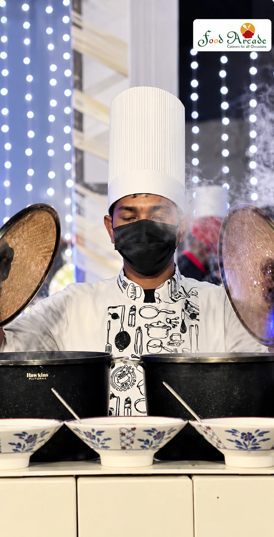 Chef in a white hat lifting lids from two steaming pots with fairy lights behind
