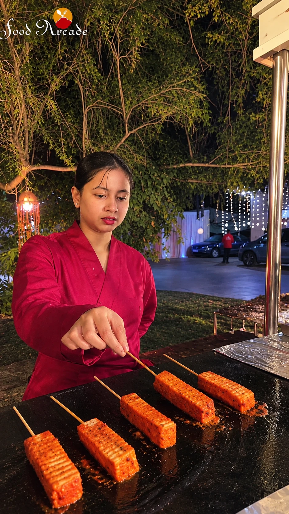 Server in a magenta jacket turning marinated paneer skewers on a griddle at a garden event at night